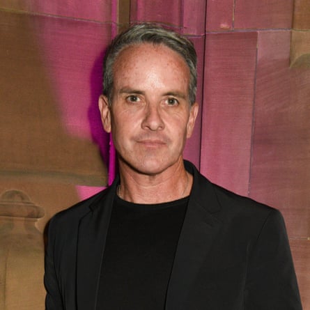 Sacha Lord, with short hair and a T-shirt under a blazer, stands against a wall looking serious