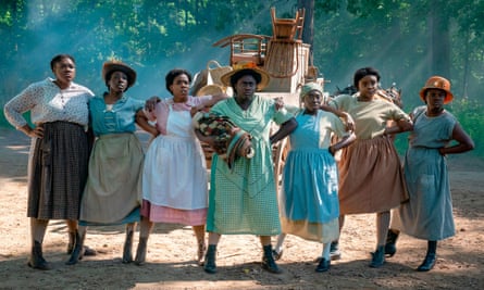 Danielle Brooks holding a baby, standing in the middle of a line of defiant looking Black women, hands on hips, in a scene from The Color Purple.