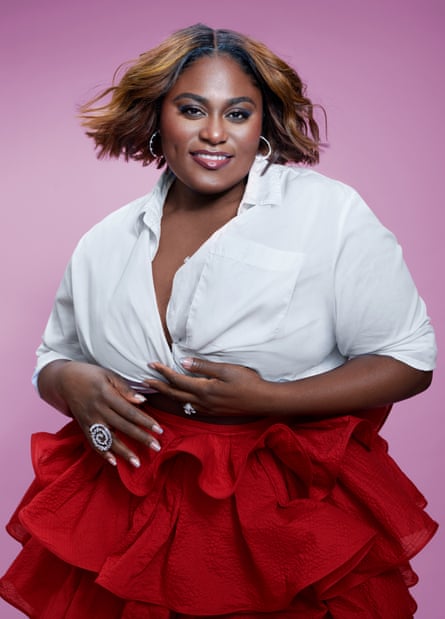 Danielle Brooks wears a red ra-ra skirt and white blouse, cinched at the waist, front buttons undone.