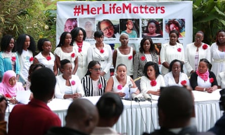 Two rows of women wearing white with a single bright red flower in front of a banner with pictures of women who have been murdered and the hashtag Her Life Matters