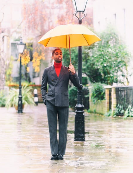 Kingsley Ben-Adir wears smart grey suit, red roll-neck jumper underneath, holding a yellow umbrella on a pretty London street in the rain
