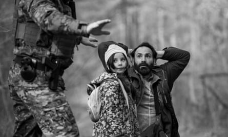 A man kneeling on the floor with a small girl standing next to him, while a soldier, cropped in the foreground, gestures at them