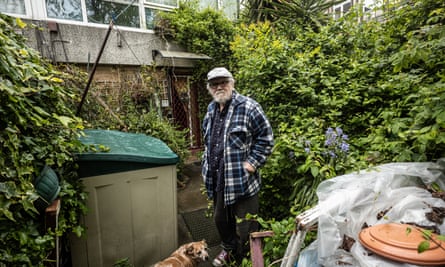 Patrick Barry in his garden; he wears a white cap, and loose checked shirt, and has a grey beard and glasses; tall bushes and yucca plants are seen in the background along with a compost or garden storage bin; and an elderly looking small brown dog stands at his feet