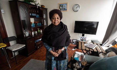 Rose Asenguah in her living room; a display cabinet with coloured drinking glasses, sofa, TV, and items including family photographs can be seen in the background