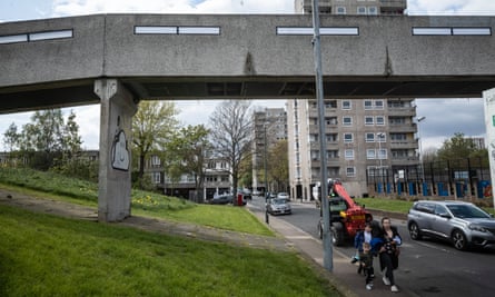 View of the estate showing concrete bridge and walkway with grassy banks, a tall tower block and a block of low-rise housing in the background; a woman walks along the pavement with two young children