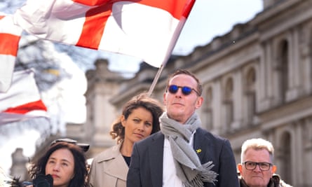 Laurence Fox standing in front of his girlfriend and people waving large flags; he is wearing dark sunglasses and a grey scarf