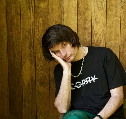 Press shot of Jonny Greenwood with his face resting in his hand, behind him wood veneer wall covering.