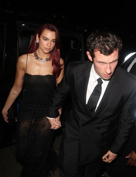 Dua Lipa in a black dress holding hands with Callum Turner in a black suit and tie, and a white shirt, outside an event at night
