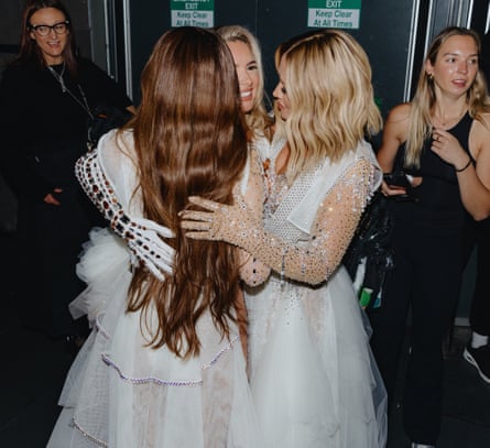 Cheryl, Nadine Coyle and Kimberley Walsh, wearing white, embellished stage costumes and having a group hug backstage at a concert, with other people in the background