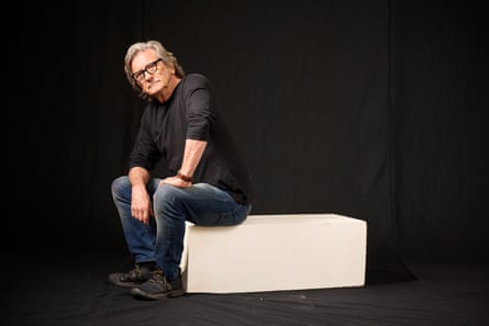 Griffin Dunne perched on the edge of a white box, against a black backdrop
