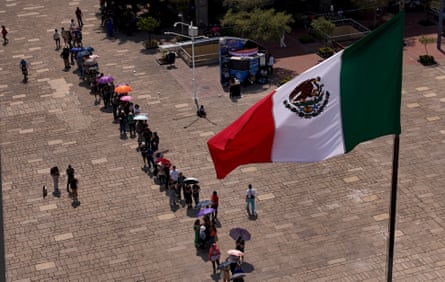 An aerial view shows a line snaking across a plaza with the Mexican flag in the foreground