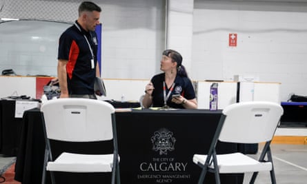 A man talks to a woman at a desk, in front of black and white chairs, with one chair back printed with: The city of Calgary Emergency Management Agency