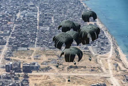 Green parachutes attached to boxes above open land, with the sea on the right hand side and dense housing at the top of the picture.