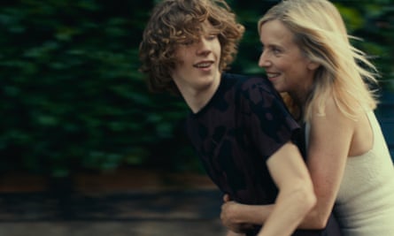 A color movie still of a young white man with an older white woman sitting behind him with her arms around his waist, as if they are on a bicycle in front of green leaves.