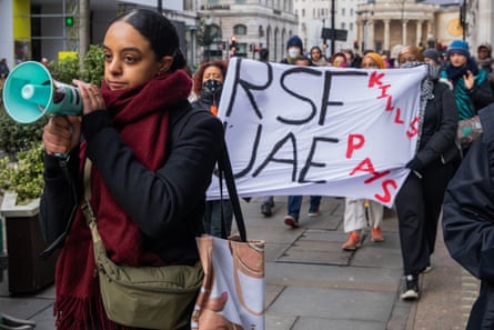 A young woman marching through the streets carrying a megaphone. A banner behind her reads: ‘RSF kills, UAE pays’