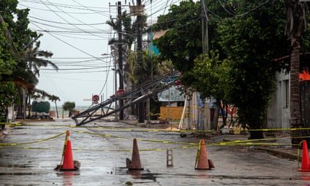 A damp urban street facing the ocean with a metal electrical tower toppled beyond orange cones.