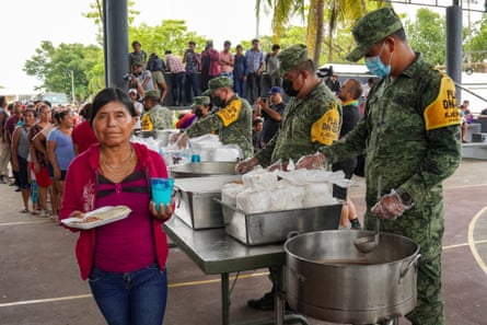 An Indigenous woman with pulled-back black hair and a red top walks away from metal tables tended by men in green camouflage, holding a plate and cup.