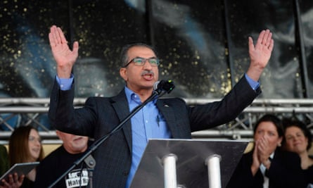 A man on a stage, speaking and holding his hands in the air