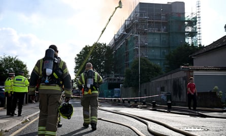 Firefighters stand in the road looking towards the block of flats in daylight. A firefighter up a turntable ladder is dousing the building with water.