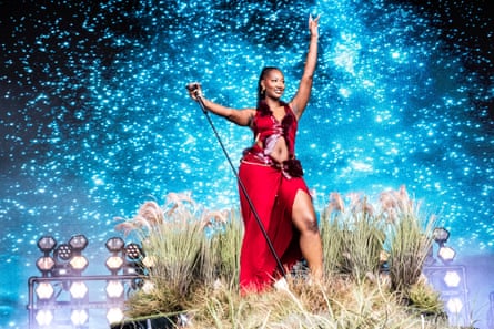 A young black African woman in a red dress stands on a tussock of grass as she performs on stage