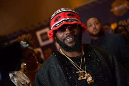 A young black African man wearing sunglasses, gold chains and a knitted hat smiles at the camera 