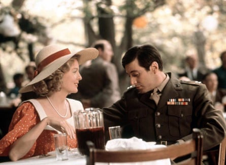 Diane Keaton wearing an old-fashioned dress and a large straw hat, and Al Pacino wearing an army uniform, sitting at a table with a large jug of red wine on it, in a garden with other people in the background