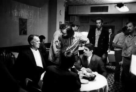 A black-and-white image of three actors in suits sitting at a restaurant table, while a director instructs them and other men stand in the background