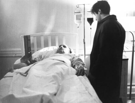 A black-and-white image of a man lying in a hospital bed, booked up to drips, while another man stands by the bed wearing a coat