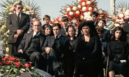 A family group, all dressed in black, sitting by a coffin, with extravagant flower arrangements behind them