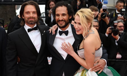 Two men in suit and ties and a woman in a green and white gown embrace each other and pose on the red carpet