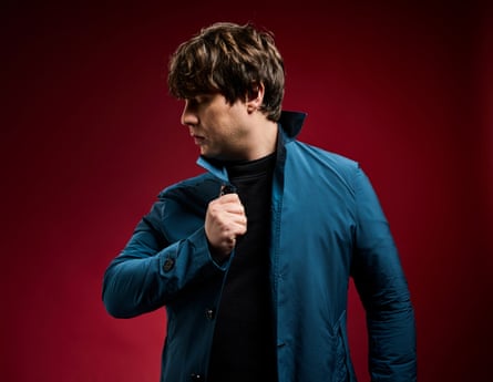 Jake Bugg in profile from waist up in blue jacket with red background in London September 2024