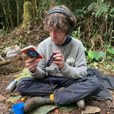 Cosmo Sheldrake is sitting on the forest floor. He is hearing headphones and holding a recording device