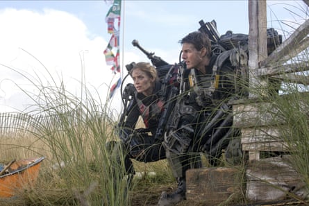 Emily Blunt and Tom Cruise wearing a mechanical battle suits, crouching beside each other in a patch of grass, with various national flags hanging from a pole in the background