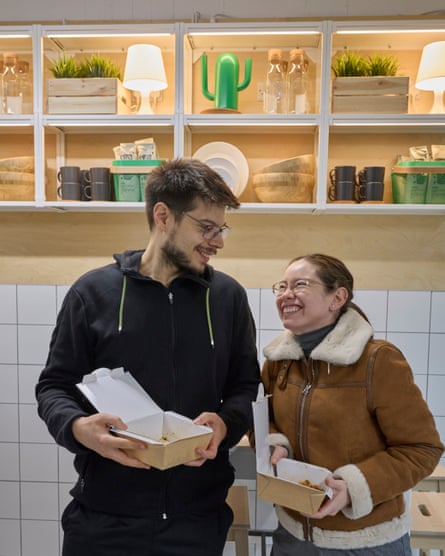 Miguel and Karen stand in front of some homewares on display in the restaurant: they are holding cardboard takeaway cartons of food and laughing