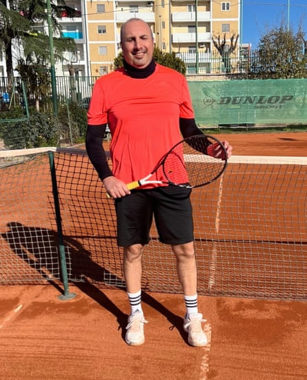 Antonio Tarantino stands holding his racket in front of the net on a red clay tennis court; he is smiling and wears a bright red T-shirt over black long-sleeved top and black shorts