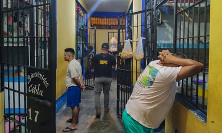 Cells are searched during an operation at the Pampas of Sanaguillo prison in Tarapoto, Peru, January 2024: two men dressed in T-shirts and shorts stand outside their cells, one with his hands behind his head and leaning against the window bars, the other with his hands behind his back. A prison guard walks down the corridor. The walls are painted bright yellow and the bars of the doors and windows are bright blue. 