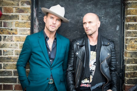 Matt in a dark turquoise suit, with Luke in motorbiker leather jacket and black T-shirt, standing against a wall.