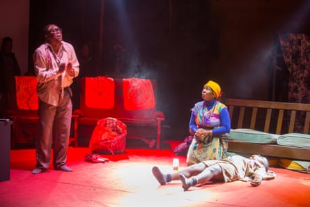 A man stands on a stage under red lighting while a woman kneels next to the stretched out body of a teenager.