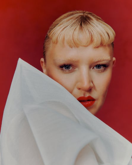 Portrait of singer Self Esteem with cropped blond hair against a red background wearing a very structured white jacket.