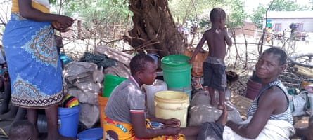 A group of people sit in the shade under a tree next to some plastic buckets and bags