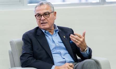 Anel Flores, in a shirt and jacket and glasses and seated in a chair, lifts his left hand as he talks