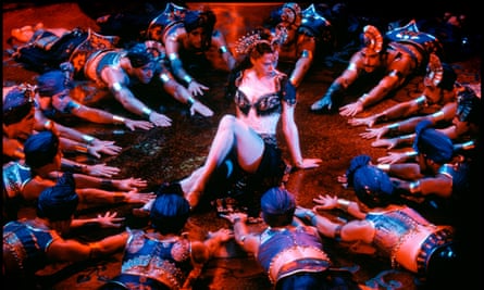 A female cabaret dancer sits on stage, at the centre of a circle of male cabaret dancers, who are lying down facing her with their arms outstretched.