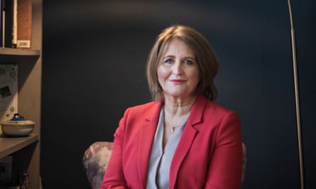 Anne Longfield at home; she sits in an armchair against a dark grey wall next to a shelf unit, and wears a red jacket.