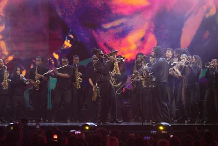 A crowd of brass musicians standing on stage playing saxophones, trumpets, flutes, a trombone and a tuba