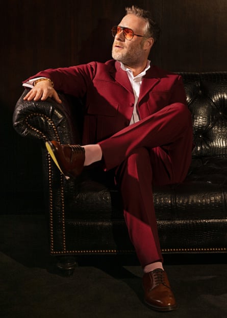 Actor Seth Rogen, in deep red suit and white shirt, sitting on leather sofa