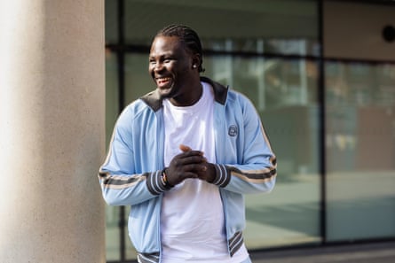 Ade Adedeji: he is smiling and holds his hands together. He wears a white T-shirt and pale blue tracksuit top.