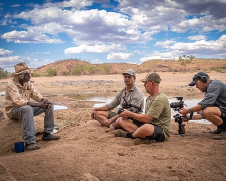 Four men sit on the dirt ground, one holding a camera, in front of mountains and under a blue sky